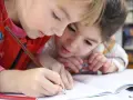 Dwie dziewczynki, jedna ma 5 latek, wspólnie rysują kredkami w zeszycie.