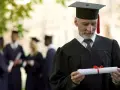 Starszy pan w todze z dyplomem zastanawia się, czy studia liczą się do stażu pracy. W tle inni absolwenci.
