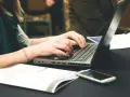 Dłonie piszą na laptopie, obok leży telefon i dokumenty. Może to być praca nad statutem szkoły.