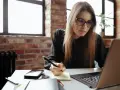 Młoda kobieta w okularach pisze notatki, przygotowując dodatek motywacyjny do CV. Na biurku laptop, telefon i notatnik.