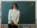 Nauczyciel początkujący z okularami trzyma stos książek przed zieloną tablicą.