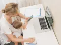 Kobieta z dzieckiem na kolanach sprawdza na telefonie informacje o zasiłku macierzyńskim 80 czy 100% i 60. Obok laptop i notatnik.