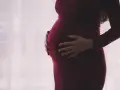 Kobieta w ciąży, z dłońmi na brzuchu, w ciemnoczerwonej sukience. Myśli o zasiłku macierzyńskim po ustaniu zatrudnienia.