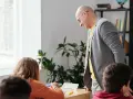 Nauczyciel pochyla się nad uczennicą, która pisze. W tle rośliny i regał z książkami. Czy to początek choroby zawodowej nauczycieli?