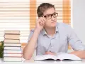 Młody mężczyzna w okularach z długopisem w dłoni zastanawia się nad treścią otwartej księgi. Obok stos książek, jakby przygotowywał się do egzaminu, który może wpłynąć na jego statut szkoły.