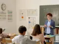 Uśmiechnięta dyrektor szkoły, w okularach, tłumaczy coś uczniom przy tablicy. W klasie wiszą kalendarze i mapy.