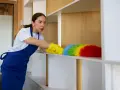Kobieta w fartuchu, niczym sumienna dozorca, czyści półkę kolorową miotełką.