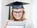 Uśmiechnięta studentka w birecie i okularach trzyma stos książek, zastanawiając się, ile trwają studia podyplomowe.