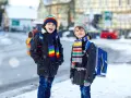 Dwóch chłopców w kolorowych czapkach i szalikach, gotowych na ferie 2027. Śnieg pada, a oni z plecakami czekają na przygodę.