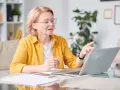 Kobieta w okularach i żółtej koszuli pracuje przy laptopie, trzymając długopis. To jej dodatek funkcyjny do pracy zdalnej.