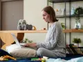Kobieta na łóżku pisze wniosek o urlop rodzicielski na laptopie, otoczona zabawkami i pluszakami.