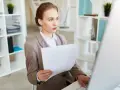 Młoda kobieta w biurze, przygotowująca się do szkolenia wstępnego BHP, analizuje dokumenty przy komputerze.