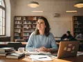Studentka z uśmiechem na twarzy, otoczona książkami i laptopem, w bibliotece. Jej przygotowanie pedagogiczne przynosi jej satysfakcję.