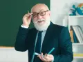 Uśmiechnięty starszy nauczyciel w okularach, z białą brodą, trzymający długopis. Myśli o zasłużonych emeryturach dla nauczycieli.