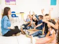 Nauczycielka siedzi na podłodze, a dzieci z podniesionymi rękami odpowiadają na pytania. Widać, że frekwencja w szkole jest wysoka, a uczniowie chętnie uczestniczą w lekcji.