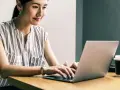 Kobieta uśmiecha się, pracując przy laptopie. Jej przygotowanie pedagogiczne online przebiega sprawnie.