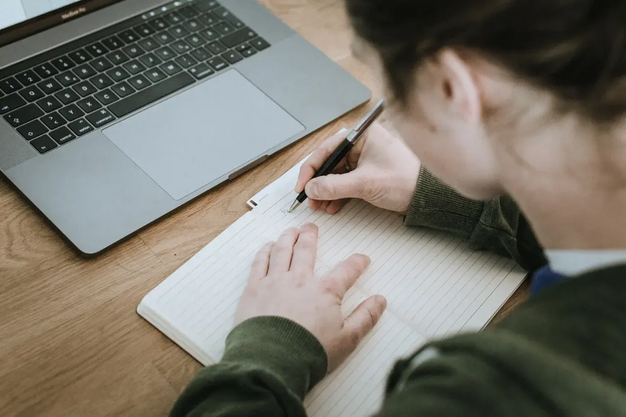 Osoba pisze w zeszycie, obok leży laptop. Powstaje opinia o uczniu, która zostanie zapisana.