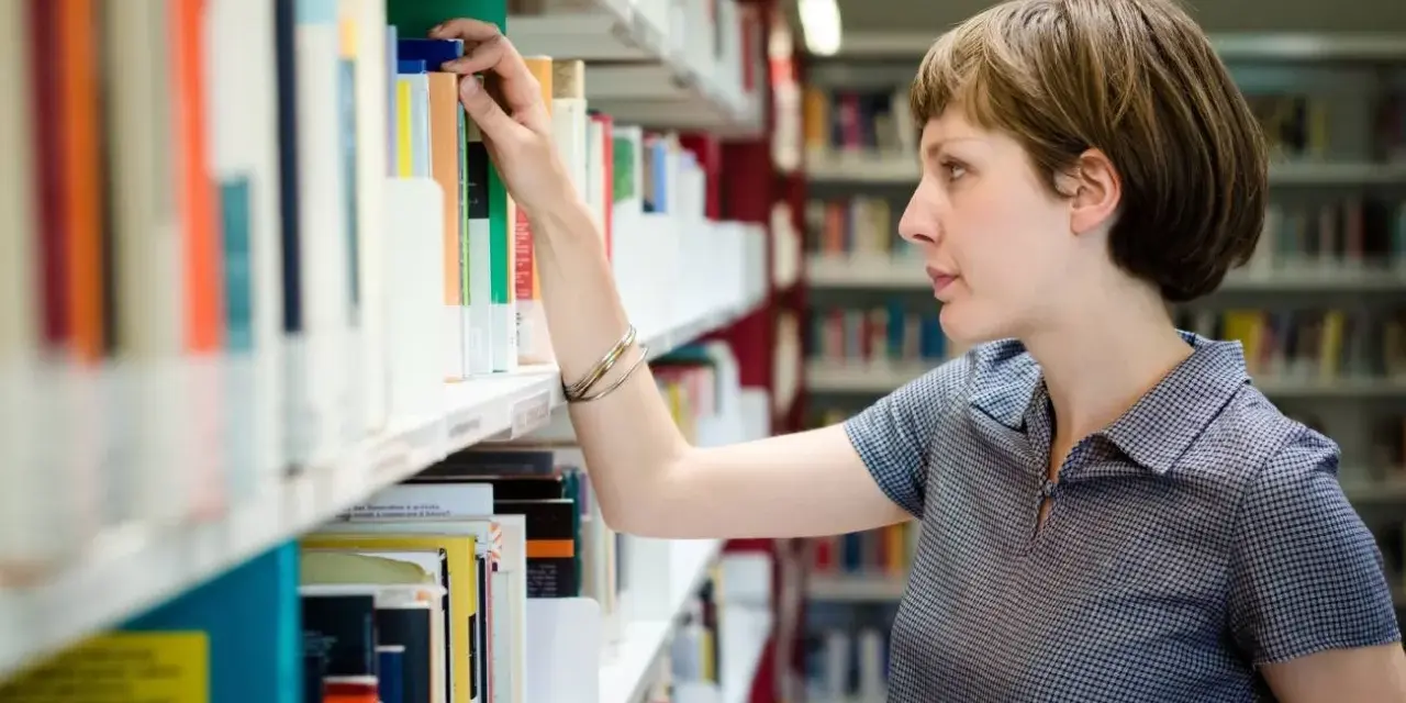 Kobieta, która wygląda jak bibliotekarz, wybiera książkę z półki w bibliotece.