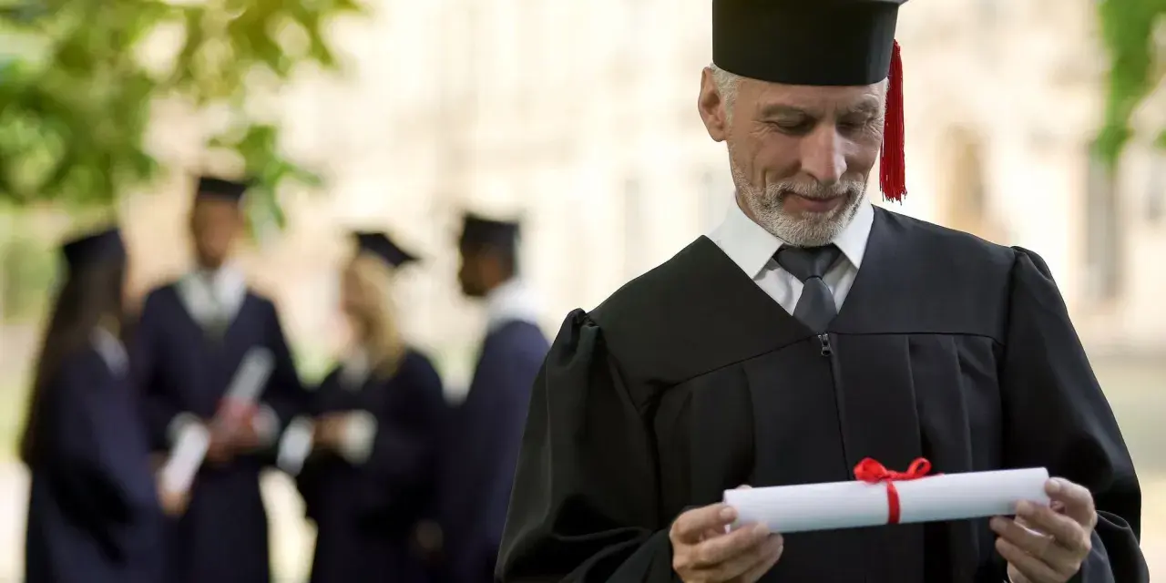 Starszy pan w todze z dyplomem zastanawia się, czy studia liczą się do stażu pracy. W tle inni absolwenci.