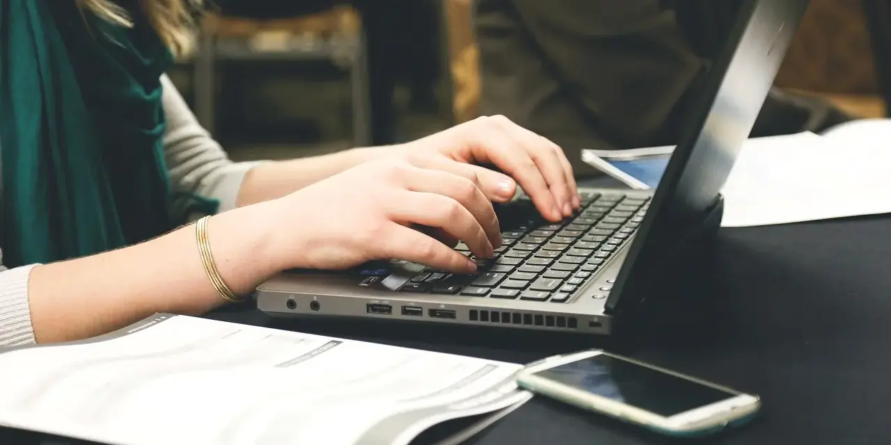 Dłonie piszą na laptopie, obok leży telefon i dokumenty. Może to być praca nad statutem szkoły.