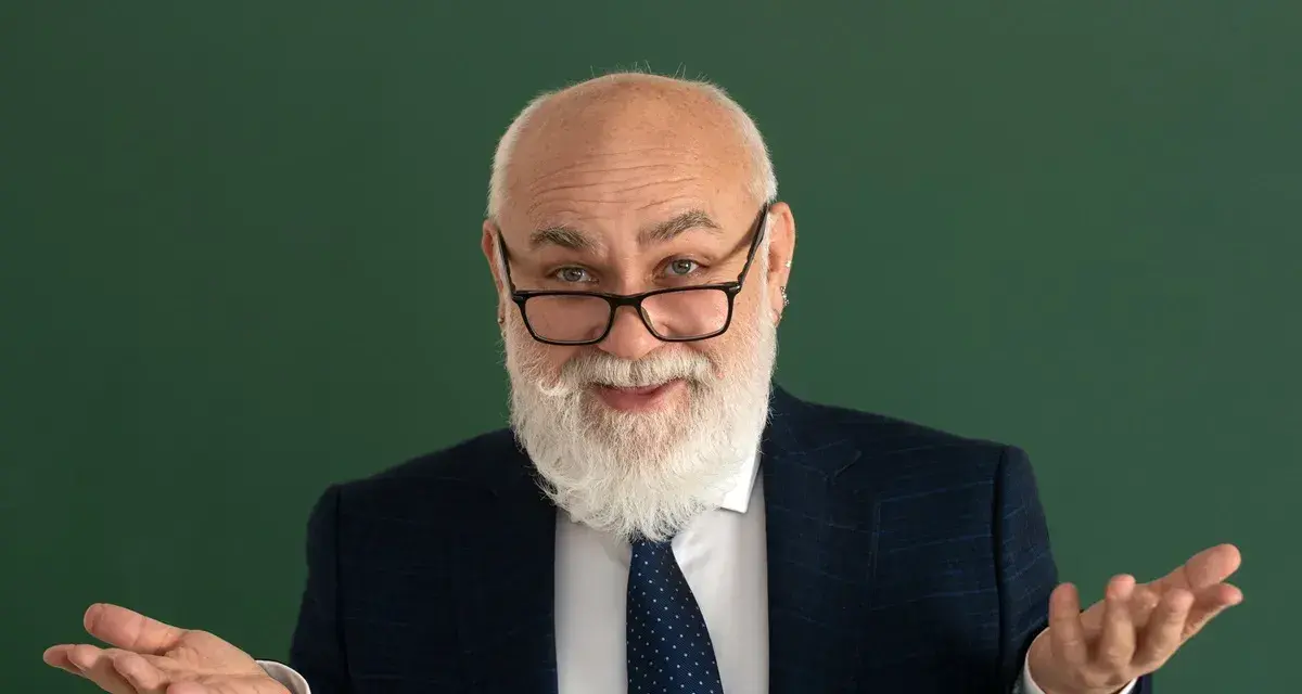 Uśmiechnięty starszy pan z brodą, w okularach i garniturze, gestykuluje, jakby wyjaśniał świadczenie kompensacyjne.