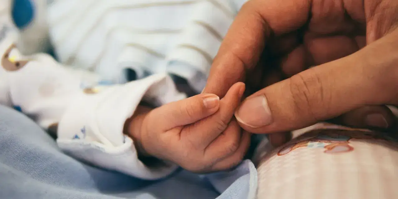 Dłoń rodzica obejmuje maleńką dłoń dziecka. To symbol miłości i troski, a także przygotowań do złożenia wniosku o urlop macierzyński.