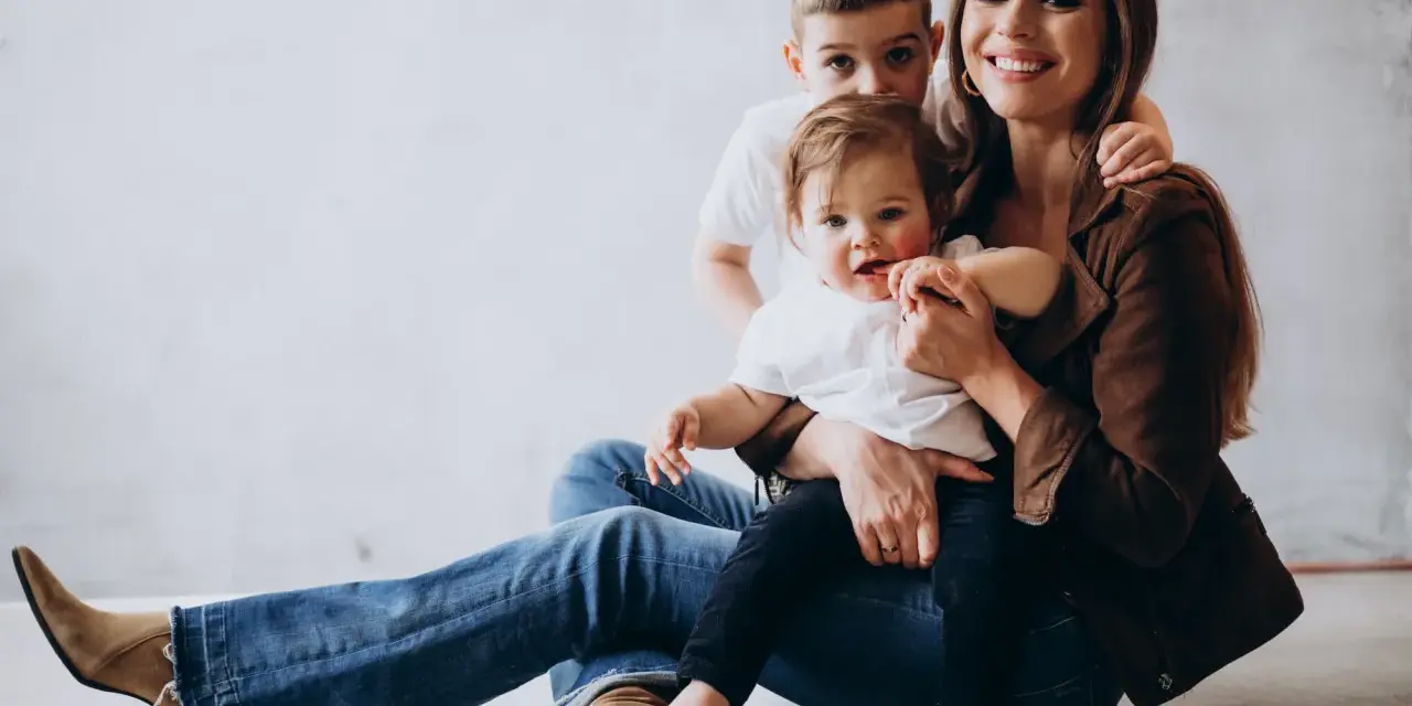 Szczęśliwa mama z dwójką dzieci, maluszkiem na rękach i starszym synkiem za plecami. Idealny urlop na dziecko.