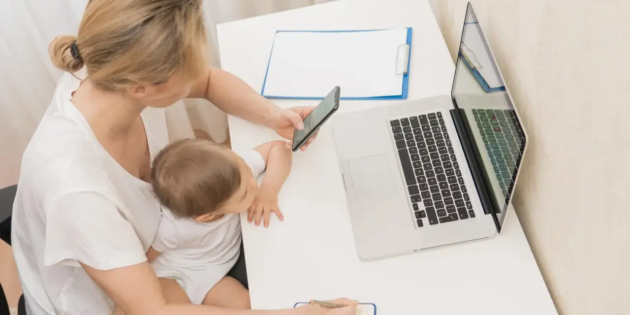 Kobieta z dzieckiem na kolanach sprawdza na telefonie informacje o zasiłku macierzyńskim 80 czy 100% i 60. Obok laptop i notatnik.
