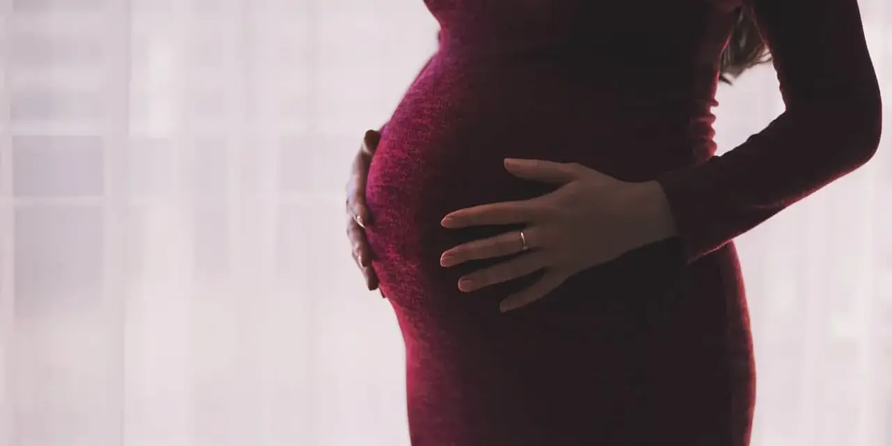 Kobieta w ciąży, z dłońmi na brzuchu, w ciemnoczerwonej sukience. Myśli o zasiłku macierzyńskim po ustaniu zatrudnienia.