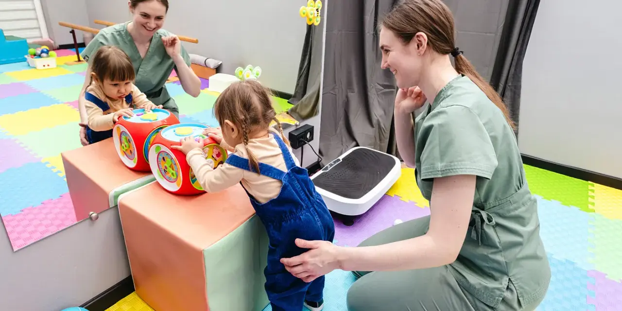 Dwie małe dziewczynki bawią się interaktywną kostką, a fizjoterapeutka wspiera ich rozwój. Www.