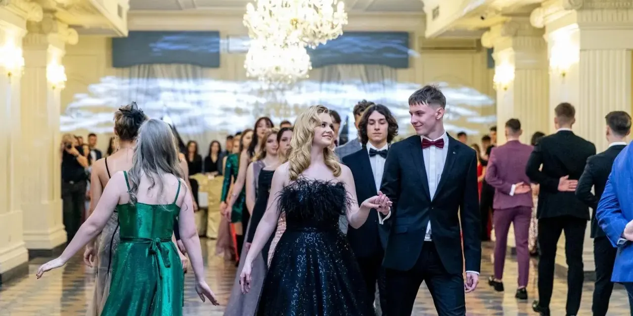 Młoda para w eleganckich strojach, ona w czarnej sukni, on w garniturze, idzie na bal studniówkowy.