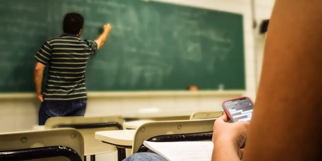 Nauczyciel pisze na tablicy, a uczeń w pierwszej ławce przegląda telefon. Obowiązki nauczyciela to nie tylko nauczanie, ale też pilnowanie porządku.
