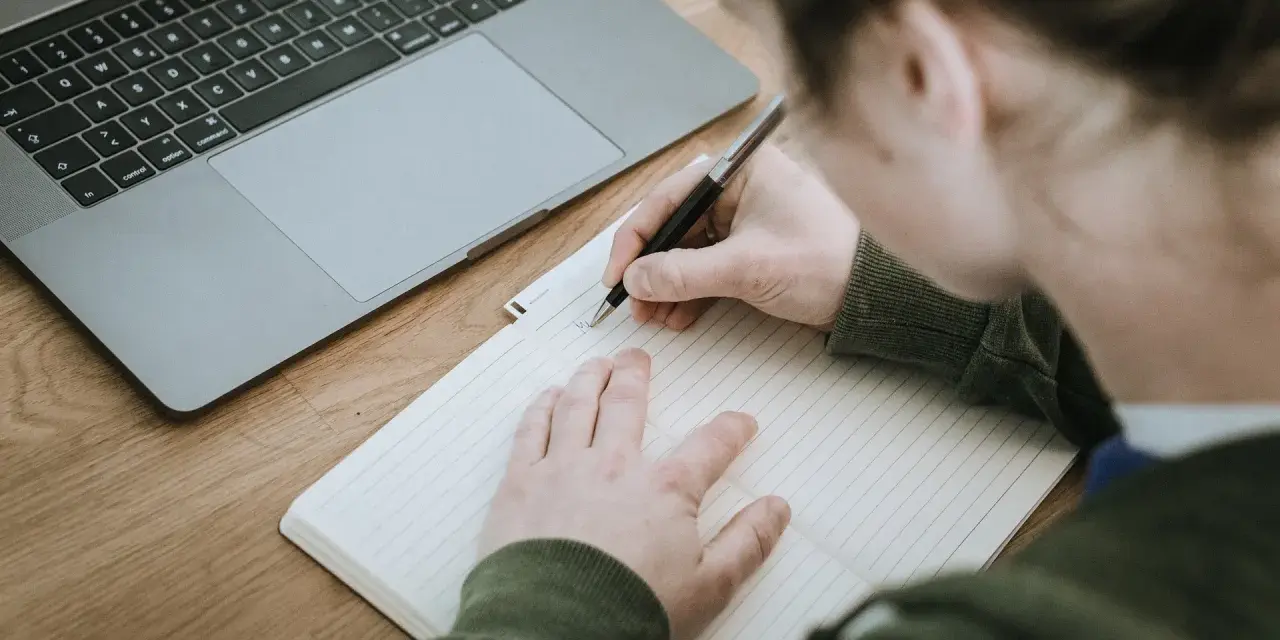 Osoba pisze w zeszycie, obok leży laptop. Powstaje opinia o uczniu, która zostanie zapisana.