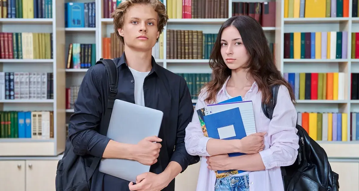 Dwóch studentów z książkami i laptopem na tle regałów z książkami.