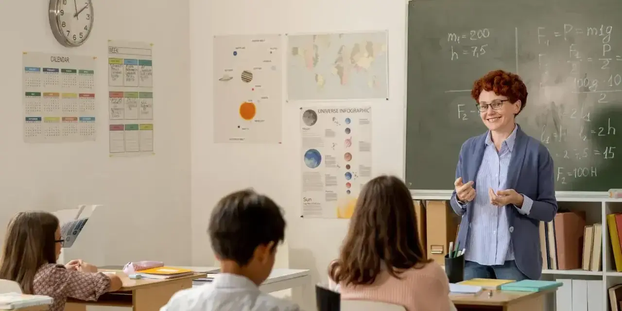 Uśmiechnięta dyrektor szkoły, w okularach, tłumaczy coś uczniom przy tablicy. W klasie wiszą kalendarze i mapy.