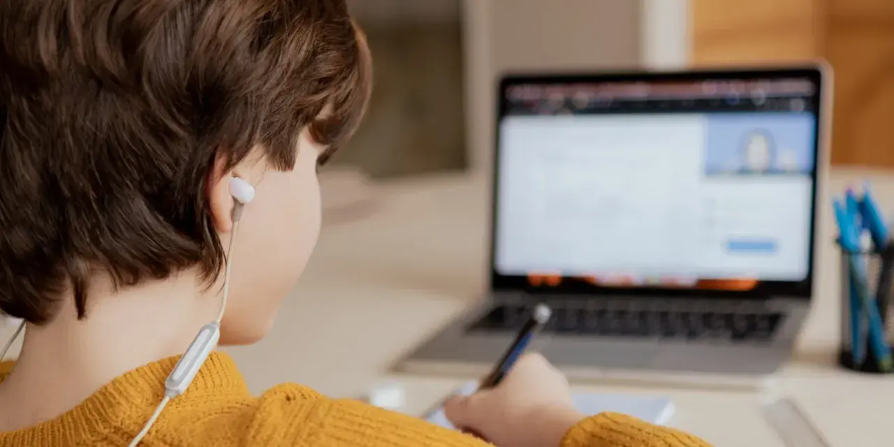 Chłopiec w żółtym swetrze uczy się zdalnie, pisząc notatki. Czy uczniowie dostaną laptopy, by móc tak pracować?