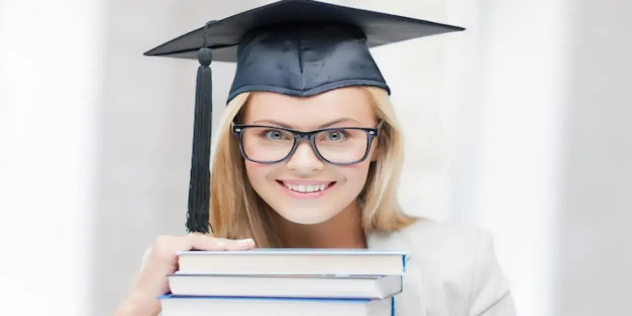 Uśmiechnięta studentka w birecie i okularach trzyma stos książek, zastanawiając się, ile trwają studia podyplomowe.