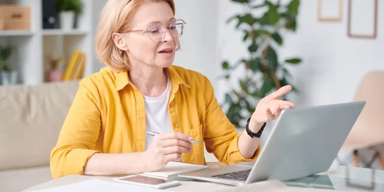 Kobieta w okularach i żółtej koszuli pracuje przy laptopie, trzymając długopis. To jej dodatek funkcyjny do pracy zdalnej.