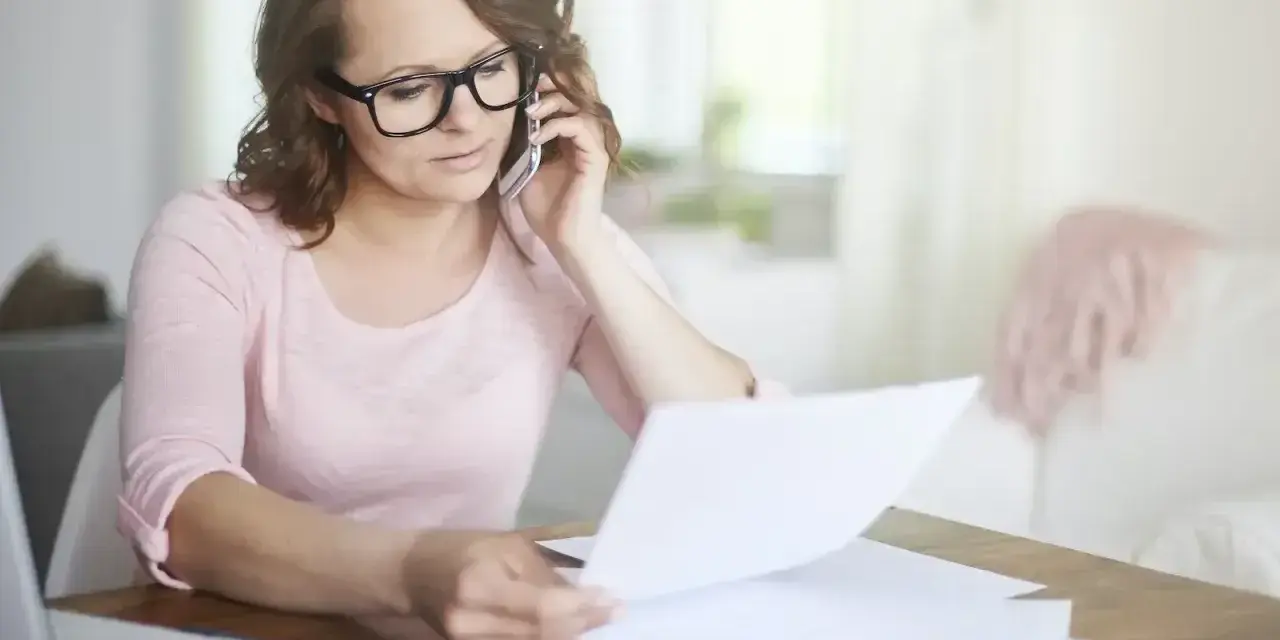 Kobieta w okularach przez telefon wyjaśnia sprostowanie świadectwa pracy, przeglądając dokumenty przy biurku.