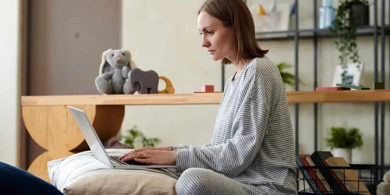 Kobieta na łóżku pisze wniosek o urlop rodzicielski na laptopie, otoczona zabawkami i pluszakami.