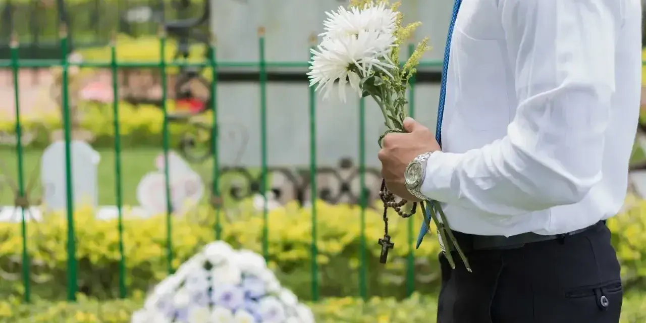 Mężczyzna w białej koszuli i niebieskim krawacie trzyma bukiet białych kwiatów i różaniec. To jego urlop okolicznościowy śmierć teścia.