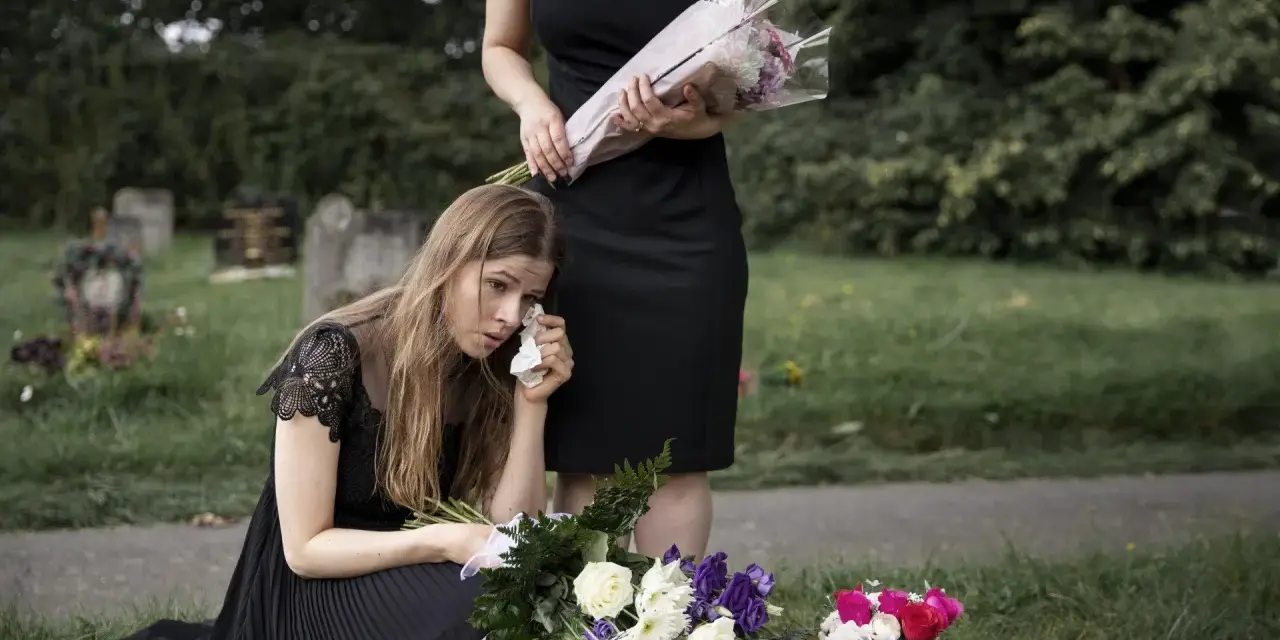 Smutna kobieta w czarnej sukience klęczy przy grobie, ocierając łzy. Obok stoi druga kobieta z bukietem kwiatów. To obraz żałoby, trudny urlop macierzyński po poronieniu.