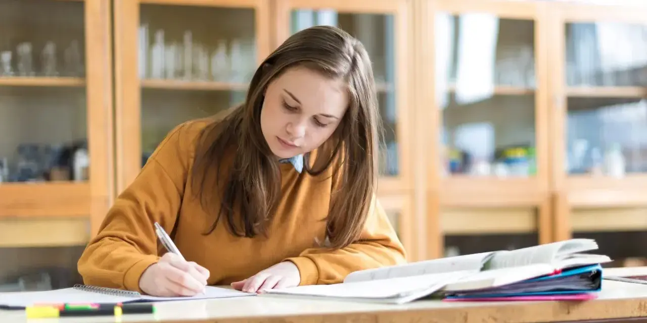 Dziewczyna pisze w zeszycie, przygotowując się do egzaminu. Marzy o stypendium szkolnym, które pomoże jej w dalszej nauce.