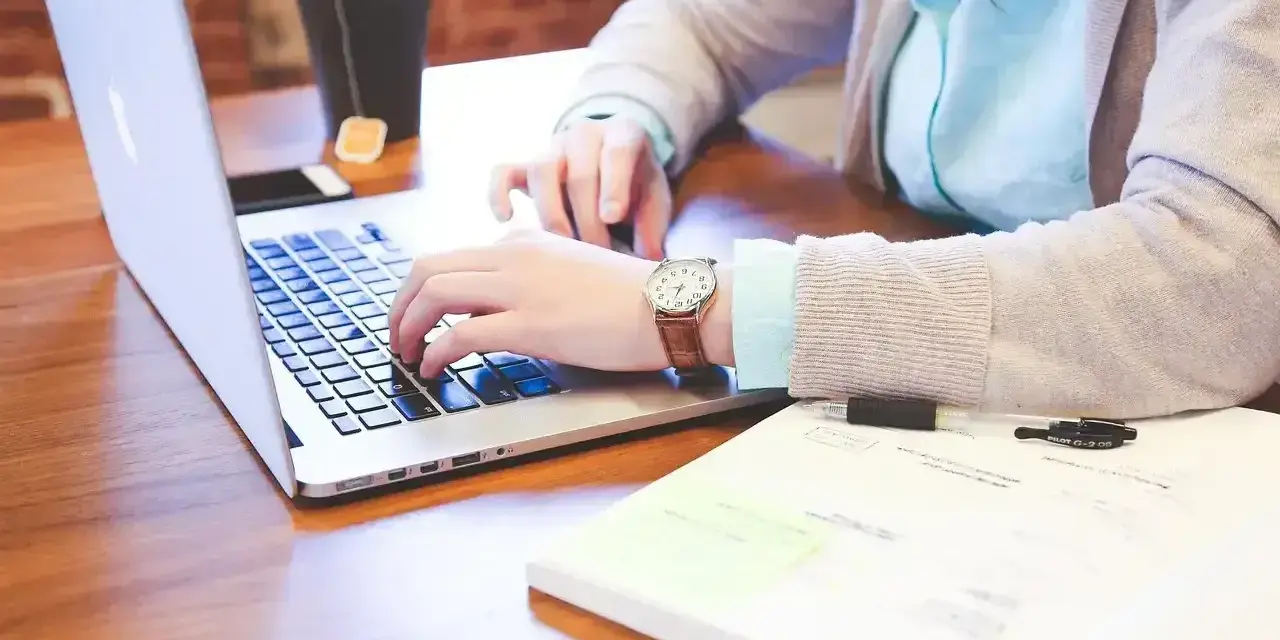 Osoba pisząca na laptopie, z kubkiem herbaty i notatkami na biurku.