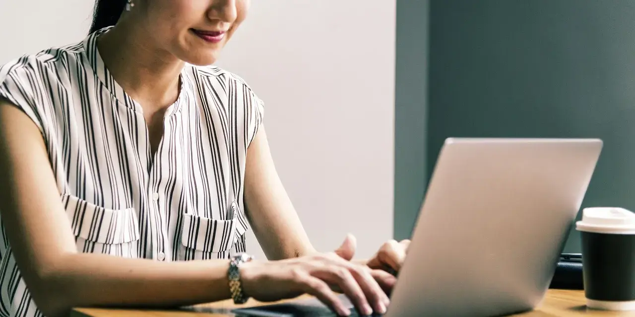 Kobieta uśmiecha się, pracując przy laptopie. Jej przygotowanie pedagogiczne online przebiega sprawnie.