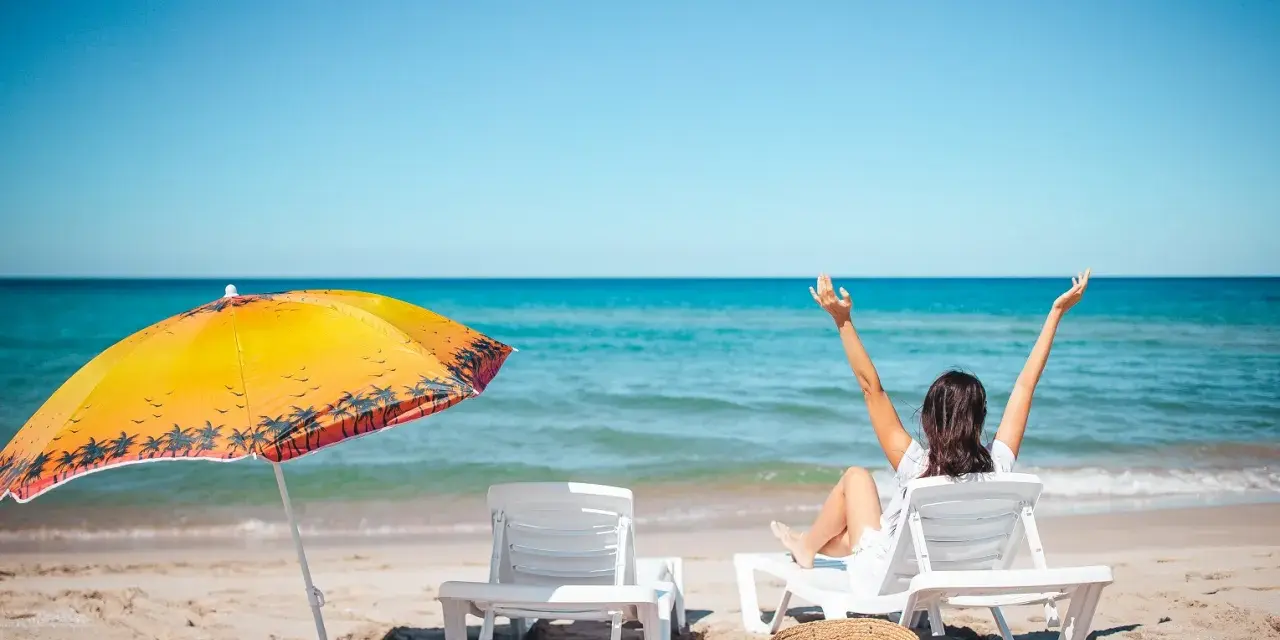 Kobieta cieszy się słońcem na plaży, w pełni wykorzystując swój urlop. Obok parasol i leżaki.
