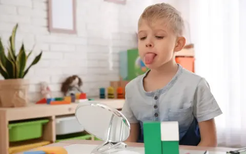 Chłopiec ćwiczy język przed lustrem, pracując nad **dyslalią**. W tle zabawki i rośliny.