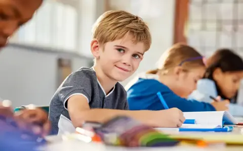 Uśmiechnięty chłopiec z blond włosami siedzi w klasie, otoczony nowymi przedmiotami w szkole. Obok niego inne dzieci piszą w zeszytach.