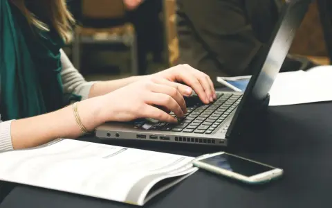Dłonie piszą na laptopie, obok leży telefon i dokumenty. Może to być praca nad statutem szkoły.