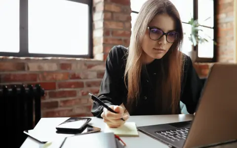 Młoda kobieta w okularach pisze notatki, przygotowując dodatek motywacyjny do CV. Na biurku laptop, telefon i notatnik.